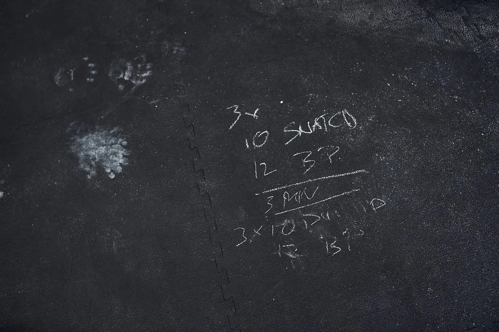 Mit Kreide auf die Bodenmatten der Halle geschriebenes Workout: 10 Snatch, 12 Burpees, 3 Minuten